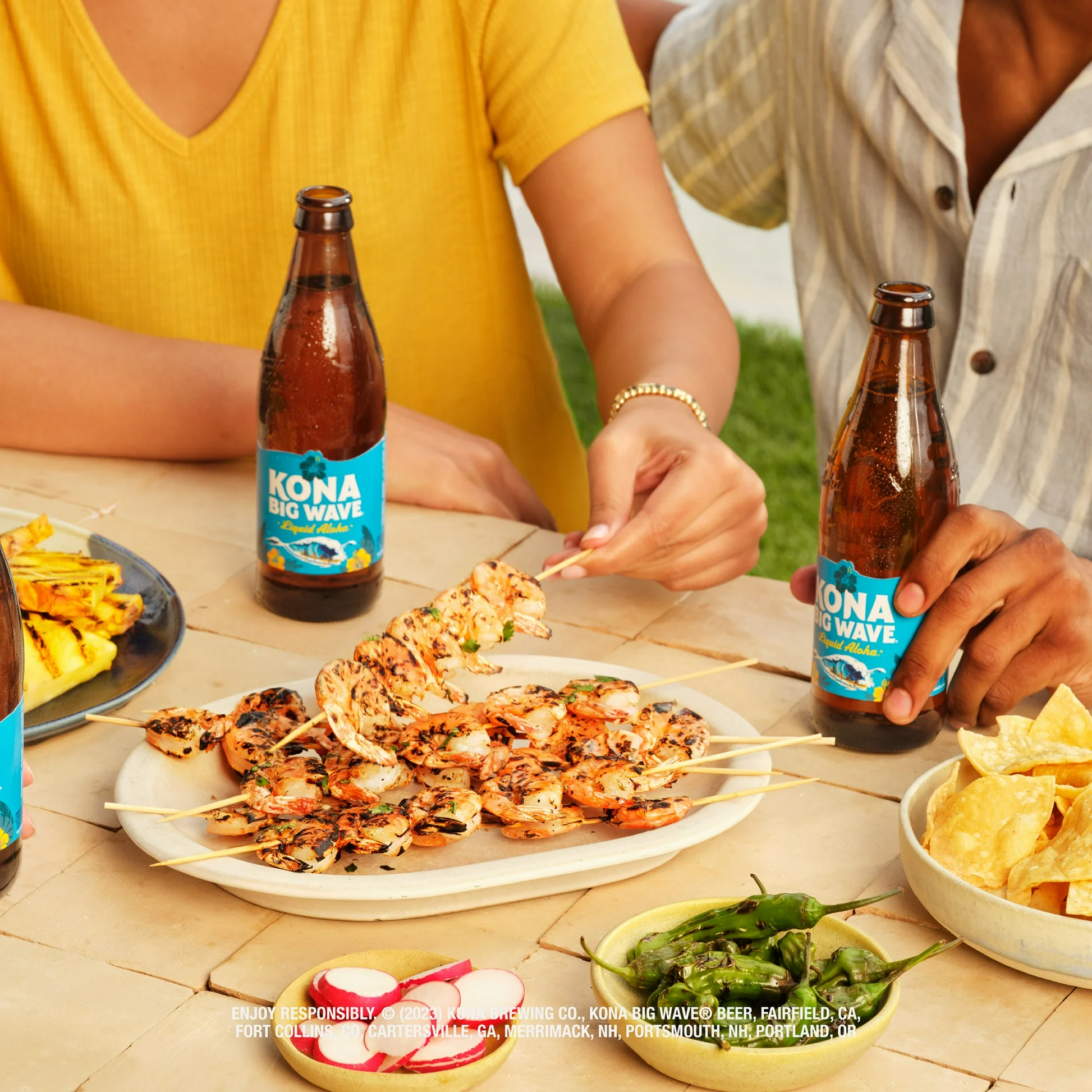 Two people enjoying grilled skewers with Kona Big Wave beer bottles on a table outdoors.