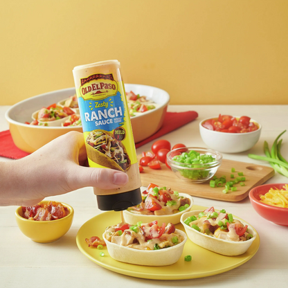 Hand holding a tube of Old El Paso Zesty Ranch Sauce over a dish with ingredients on a yellow background